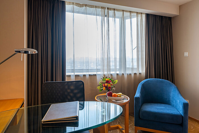 酒店客房內明亮的座位區與工作空間，配備玻璃書桌、藍色扶手椅、鮮花裝飾，並透過大窗戶引入自然光線