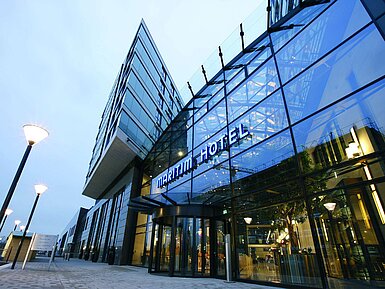 Modern exterior view of the Maritim Hotel Düsseldorf with glass facade and illuminated hotel logo at dusk.