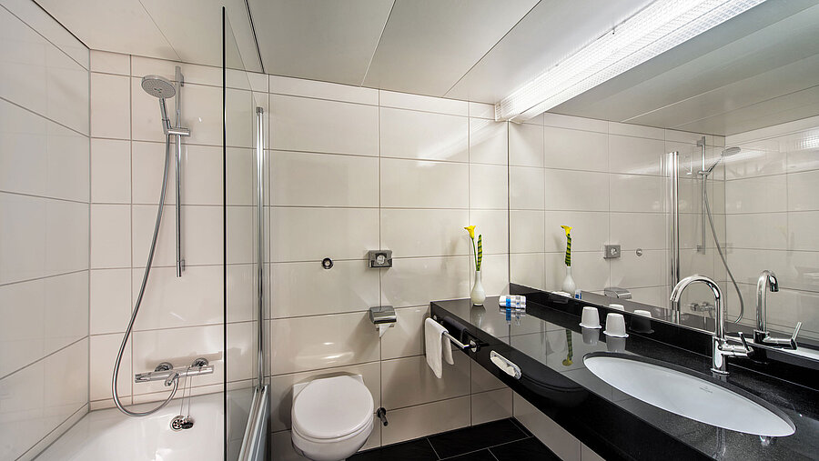 Bathroom at the Maritim Hotel Kiel with bathtub, glass shower screen, washbasin and large mirror on a black shelf.