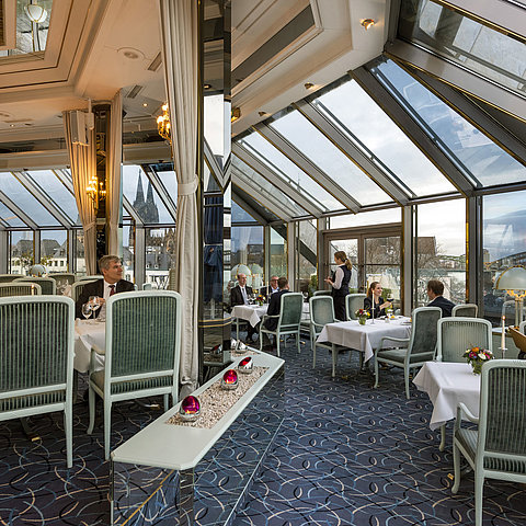 Elegant restaurant with a glass roof offering views of Cologne Cathedral and Hohenzollern Bridge, guests enjoying fine dining.