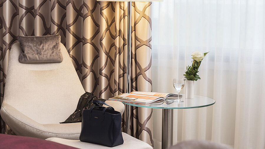 Detail of a Classic room at Maritim Hotel Bonn with a cozy chair, curtain, table, glass, rose, and a handbag.