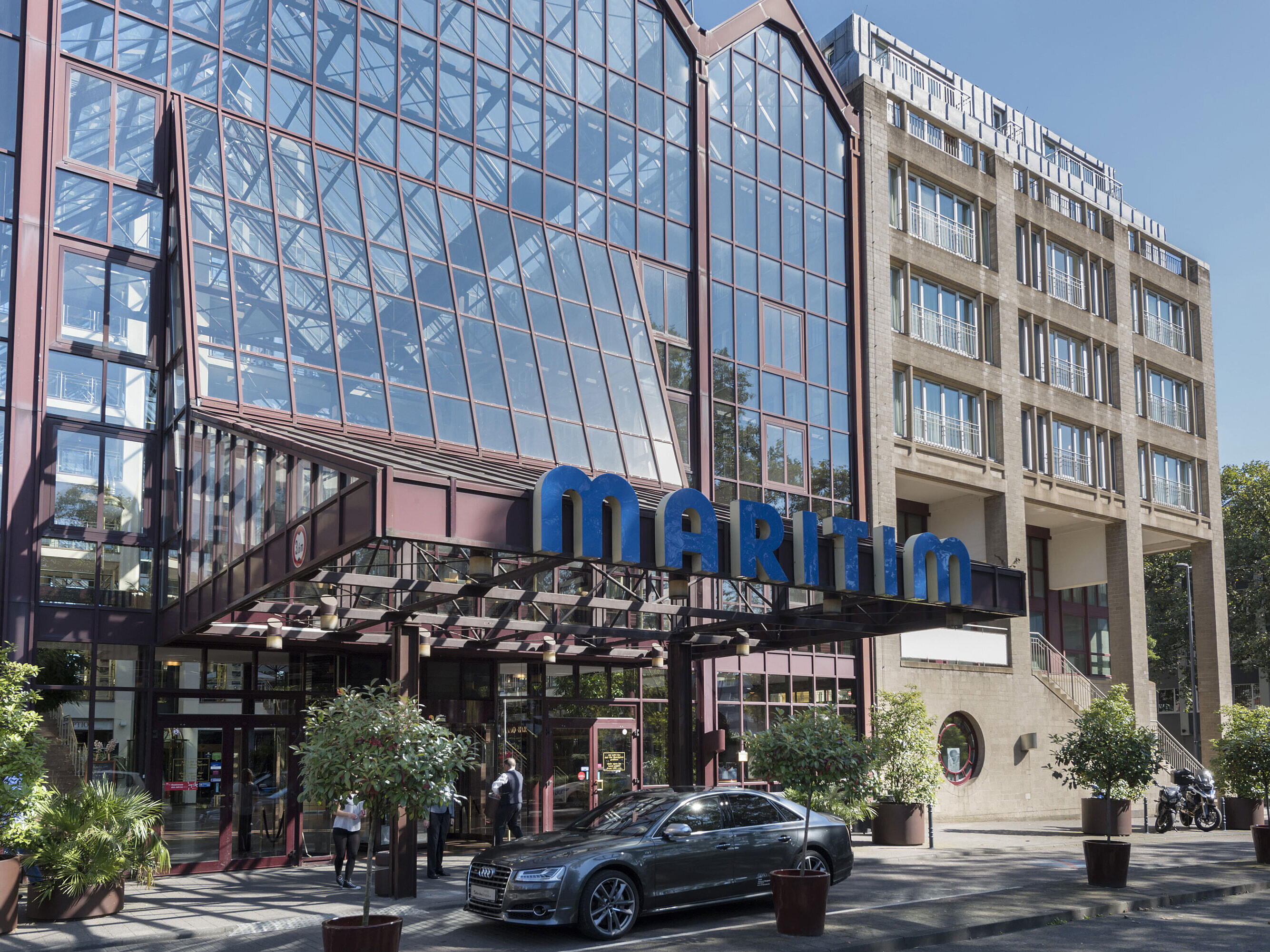 Glass facade of Maritim Hotel Cologne in daylight with Maritim sign and parked car in front of the entrance.