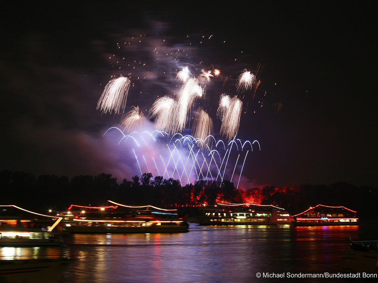 Fireworks lighting up the night sky above a river with illuminated boats
