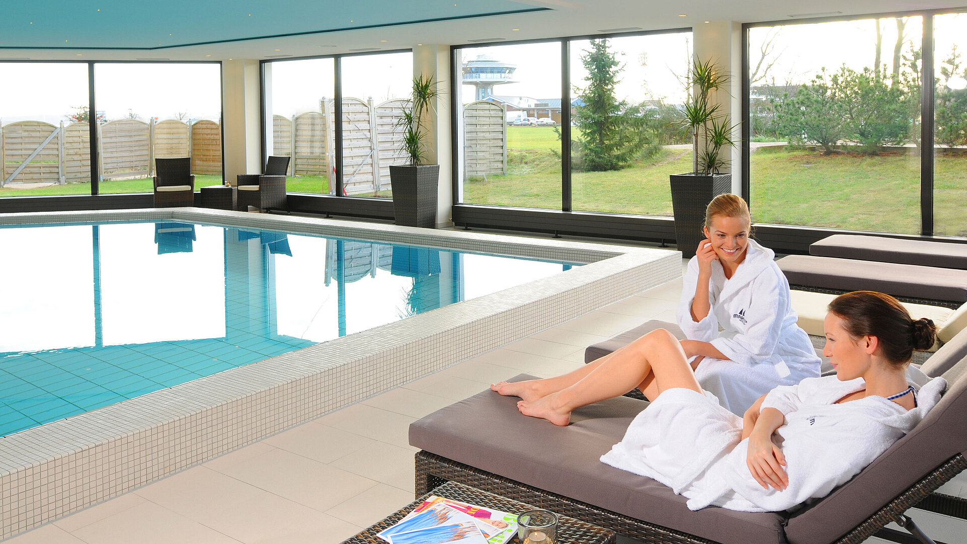 Two women relax in bathrobes on loungers at the indoor pool of Maritim Hotel Travemünde
