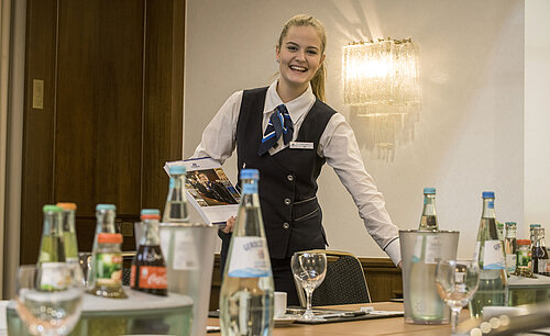 Friendly service employee in the Salon Lenné of the Maritim Hotel Bonn with drinks and conference documents on the table.