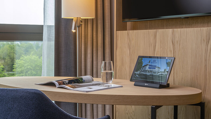 Desk at Maritim Hotel Ingolstadt with a tablet, magazine, and water glass, overlooking nature through large windows.