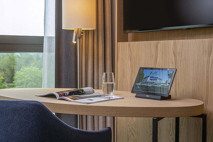 Desk at Maritim Hotel Ingolstadt with a tablet, magazine, and water glass, overlooking nature through large windows.