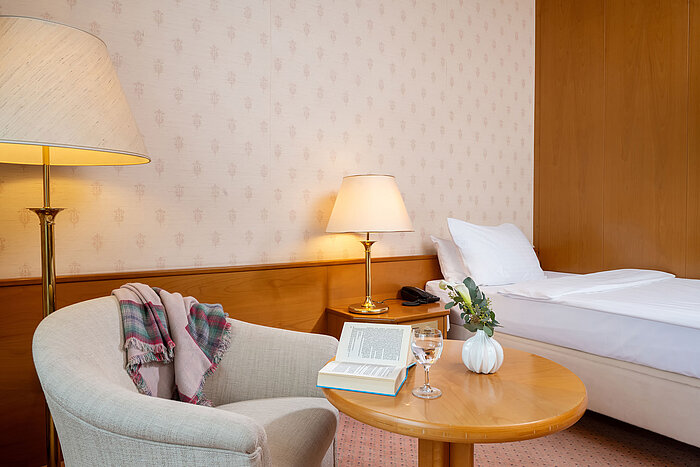 Cosy seating area with armchair, table, lamp and a view of the single bed in the hotel room.