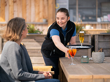 Waitress serves Aperol Spritz and beer to a guest on the terrace of the Beach Lounge, with the beach in the background.