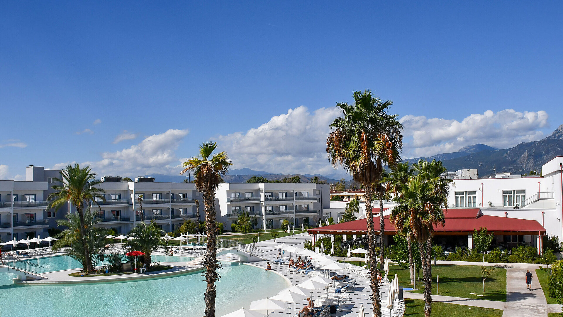 Panorama of Maritim Resort Calabria with pool, palm trees, white buildings, and mountain backdrop under clear skies.