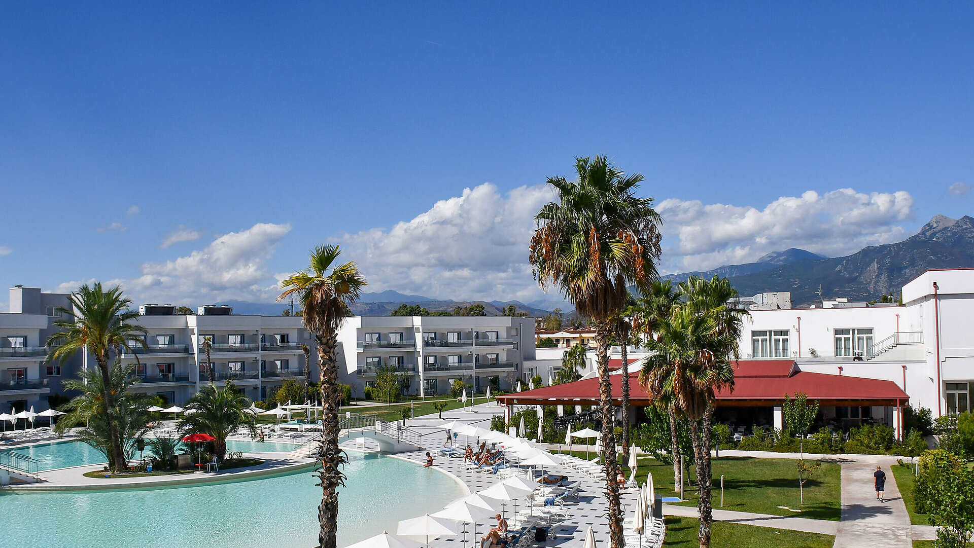 Panorama of Maritim Resort Calabria with pool, palm trees, white buildings, and mountain backdrop under clear skies.