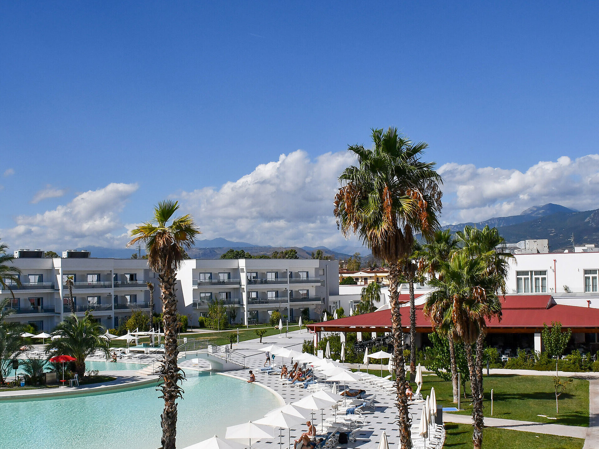 Panorama of Maritim Resort Calabria with pool, palm trees, white buildings, and mountain backdrop under clear skies.