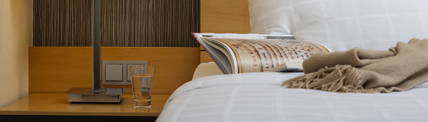 Close-up of a bedside table at Maritim Hotel Düsseldorf with a lamp, glass of water, and book on a bed.