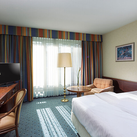 Bright Comfort room at Maritim Hotel Cologne with single bed, desk, TV, armchair, and large windows with curtains.