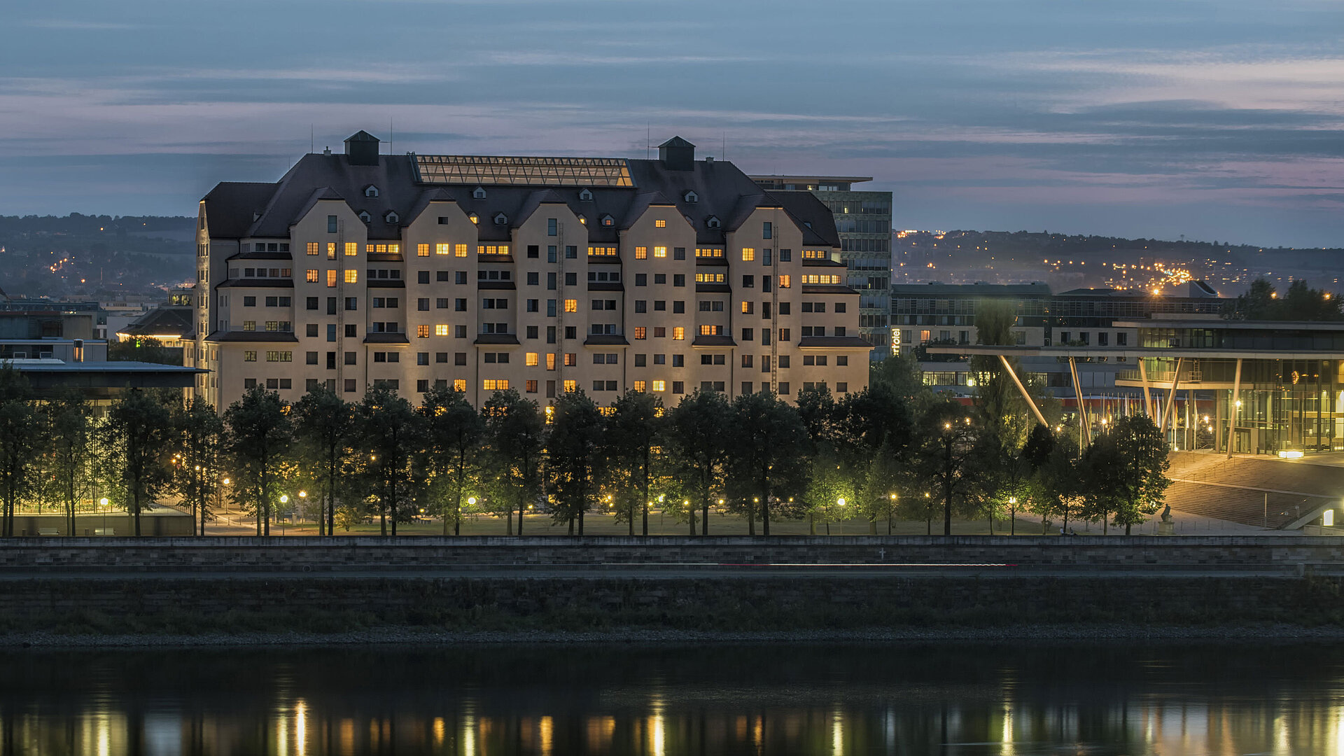 Illuminated Maritim Hotel Dresden at dusk with lights reflecting on the Elbe River.