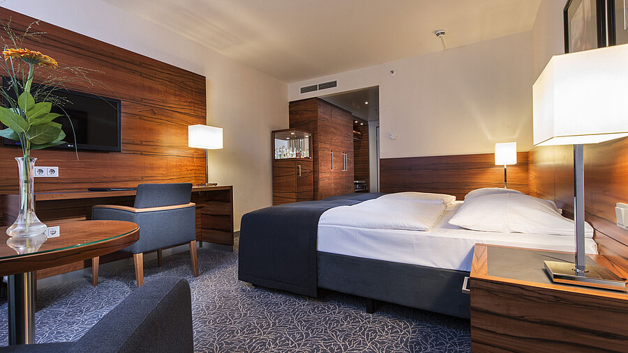 Furnished hotel room at the Maritim Hotel Munich with a comfortable bed, workspace, TV, and wood paneling.