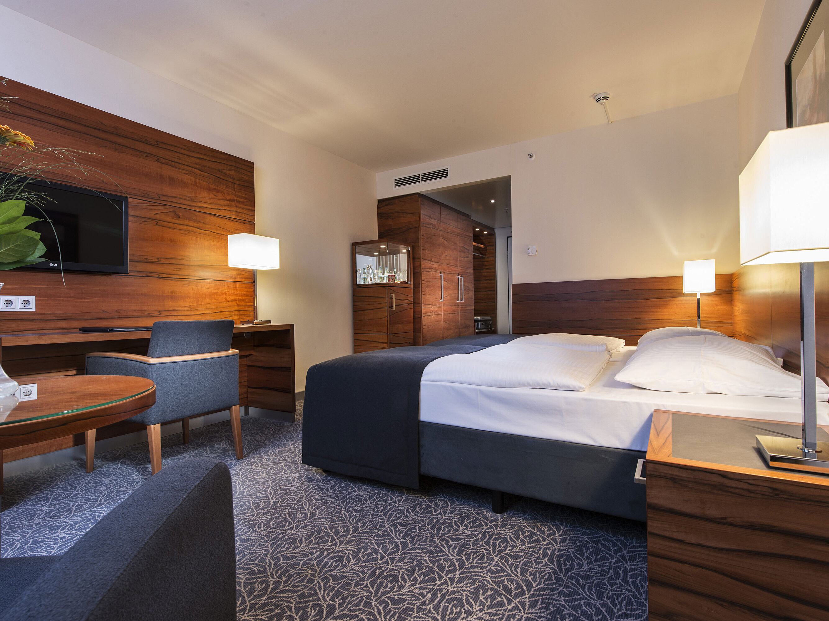Furnished hotel room at the Maritim Hotel Munich with a comfortable bed, workspace, TV, and wood paneling.