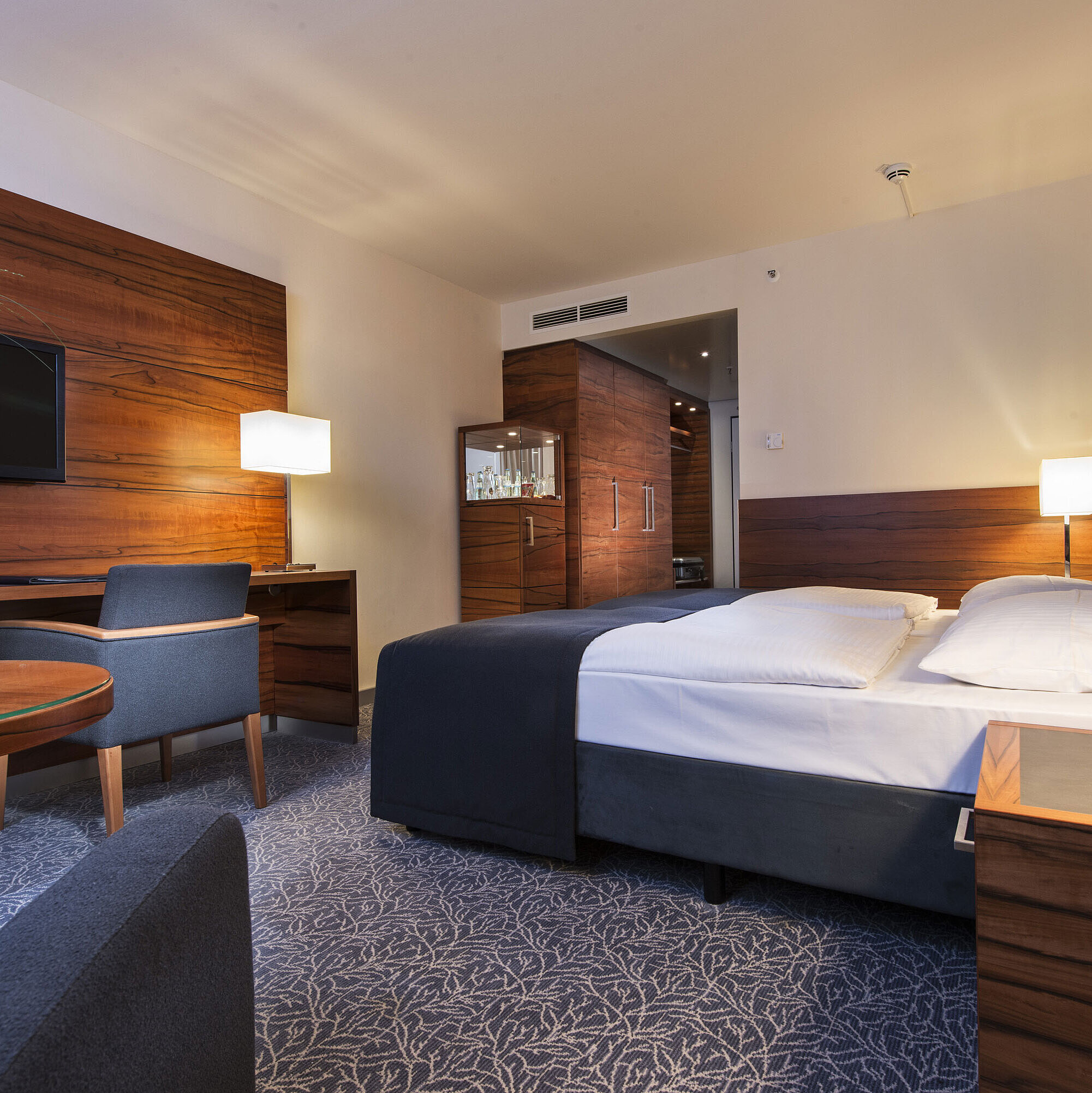 Furnished hotel room at the Maritim Hotel Munich with a comfortable bed, workspace, TV, and wood paneling.