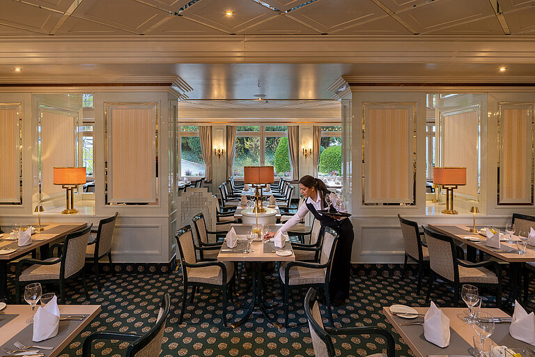Elegant restaurant with set tables, large windows and a staff member preparing the dining area.