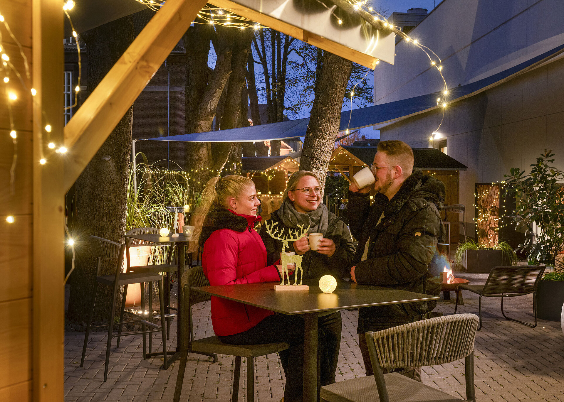 Three people enjoying hot drinks at the festive Christmas market of Maritim Hotel Darmstadt with fairy lights.