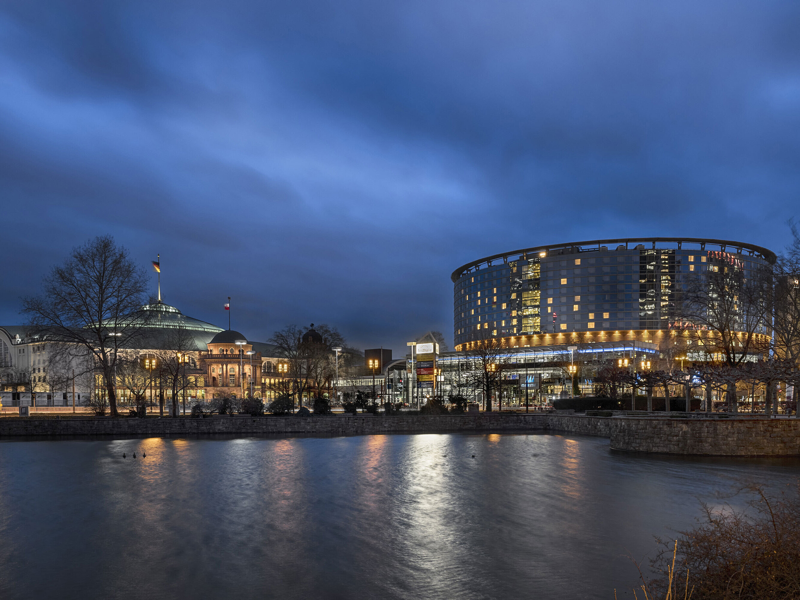 Maritim Hotel Frankfurt and Festhalle at night with illuminated glass facade and reflection in the adjacent pond.