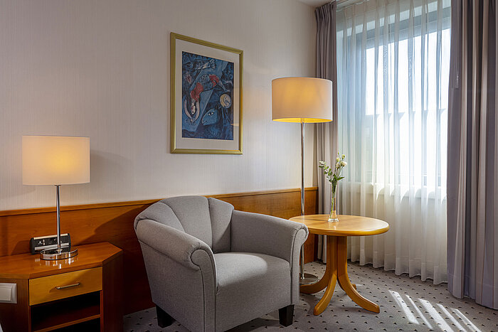 Bright hotel room in the Maritim Airport Hotel Hannover with armchair, lamp, window, and artwork.
