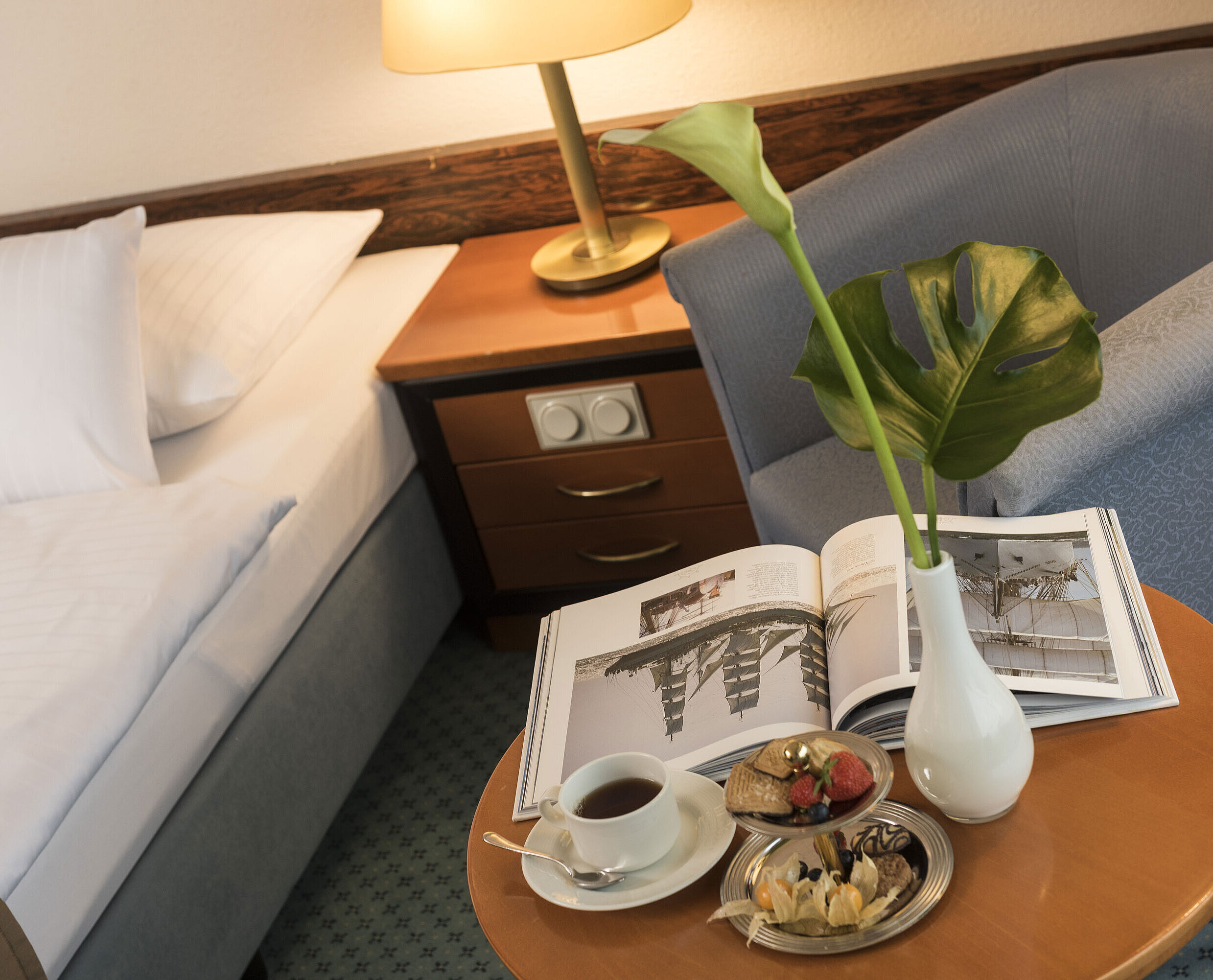 Details of a room at the Maritim Hotel Kiel: side table with vase, book and snacks next to a bed and armchair.