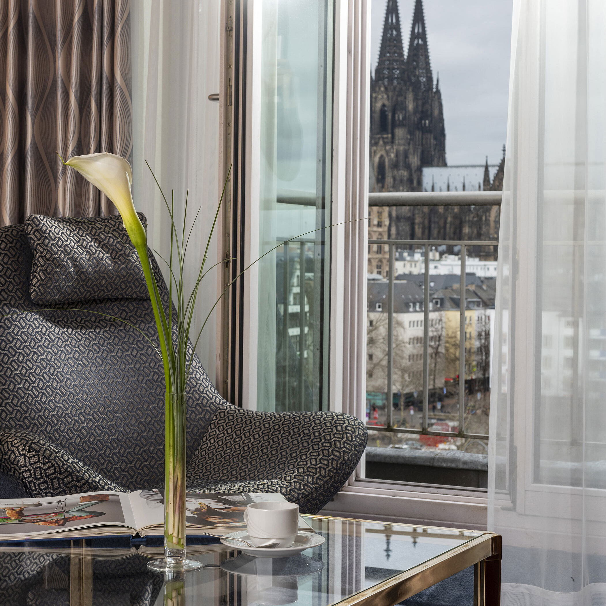 Junior Suite at Maritim Hotel Cologne with balcony view of Cologne Cathedral, modern chair, and coffee table with decoration.