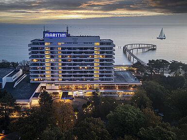 Evening view of Maritim Seehotel Timmendorfer Strand with illuminated pier and a sailboat in the background at sunset.