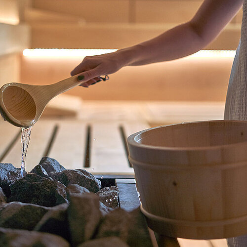 Person pouring water from wooden ladle onto hot sauna stones for infusion