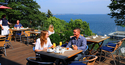 Panorama terrace at the Maritim Hotel Kiel with guests enjoying breakfast, a view of the fjord, sunny weather and a relaxed atmosphere.
