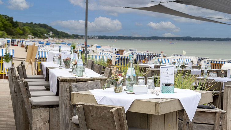Rustic wooden tables in a beach lounge, set with water bottles and glasses, with beach chairs and the sea in the background.
