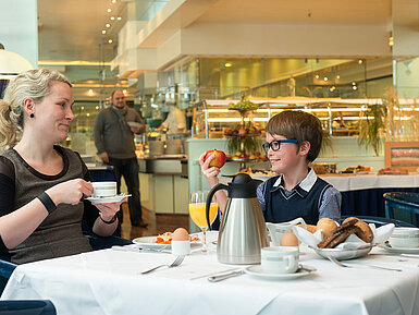 A mother and her son enjoy a relaxed breakfast at Maritim Hotel Magdeburg, with the buffet in the background.