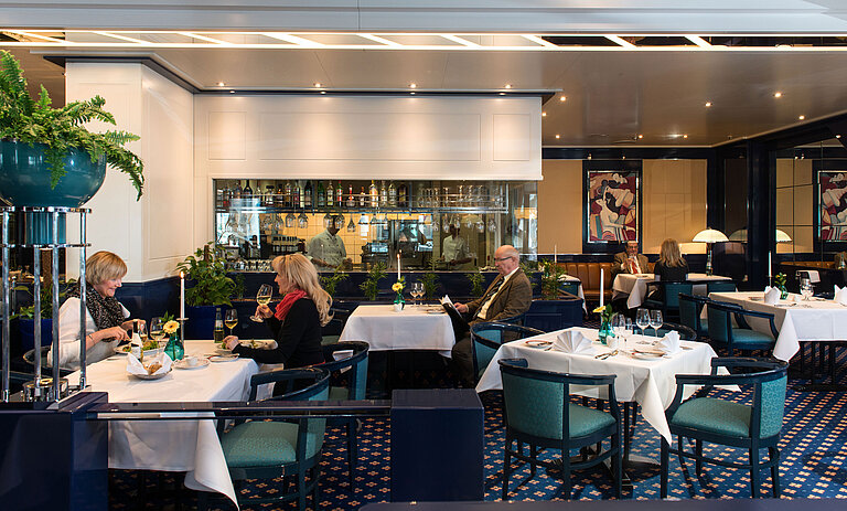 Two guests enjoying lunch in the restaurant of Maritim Hotel Magdeburg, with a view of the bar and kitchen in the background.