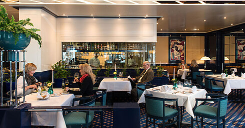 Two guests enjoying lunch in the restaurant of Maritim Hotel Magdeburg, with a view of the bar and kitchen in the background.