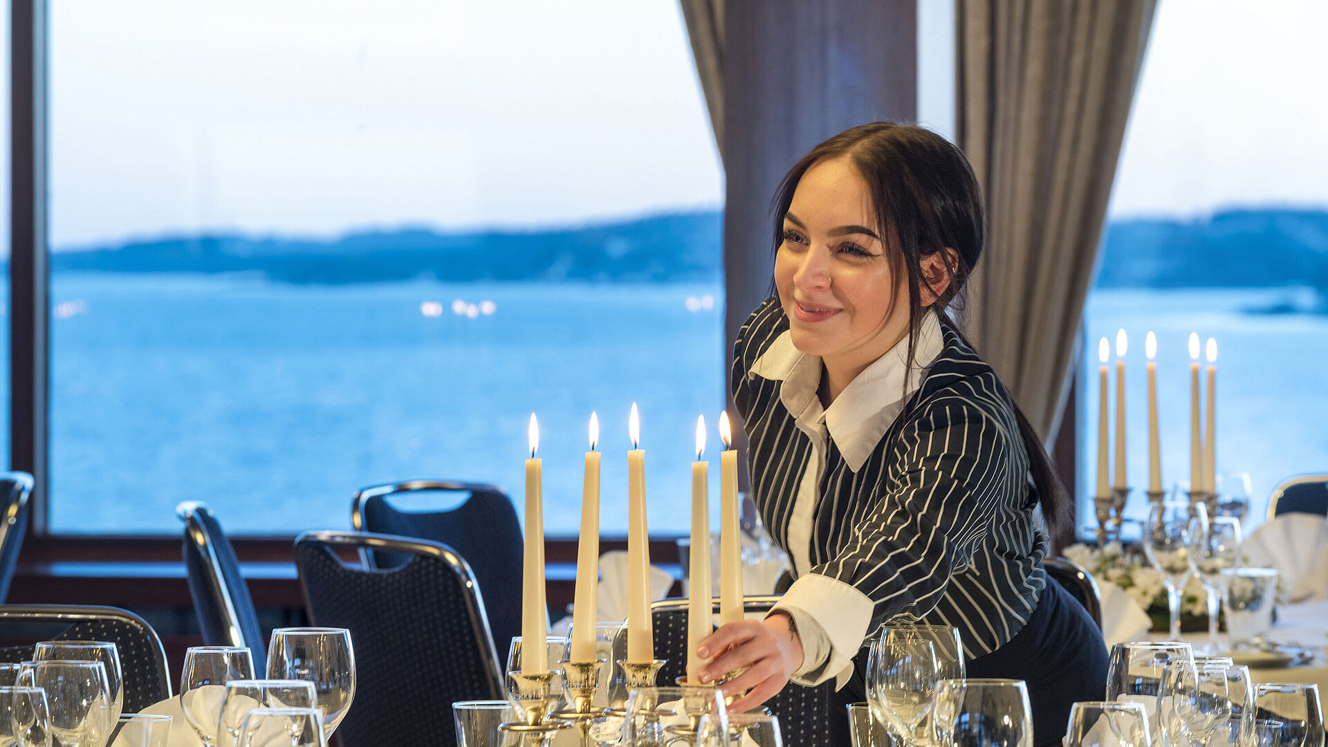 Hall Maritim in the Maritim Hotel Kiel, a member of staff lights candles on a festively laid table, sea view in the background.