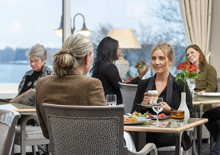 Guests in the Bellevue restaurant at the Maritim Hotel Kiel, relaxed conversation over drinks and food with a view of the sea.