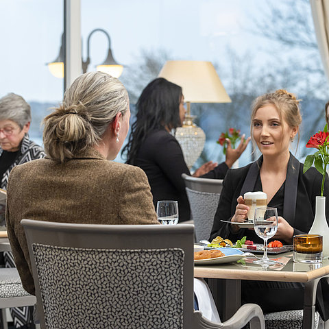 Guests in the Bellevue restaurant at the Maritim Hotel Kiel, relaxed conversation over drinks and food with a view of the sea.