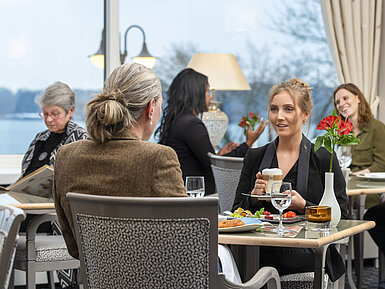 Guests in the Bellevue restaurant at the Maritim Hotel Kiel, relaxed conversation over drinks and food with a view of the sea.