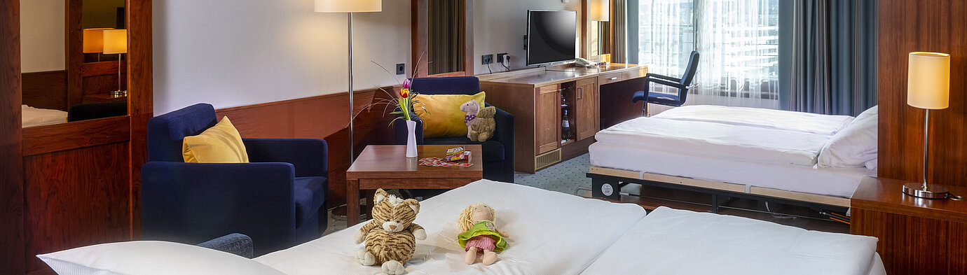 Family room at Maritim Hotel Darmstadt with toys on the bed, a desk with TV, and a cozy seating area.