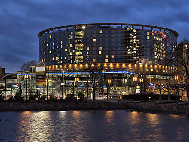 Maritim Hotel Frankfurt at night with illuminated glass facade and reflection in the calm pond.