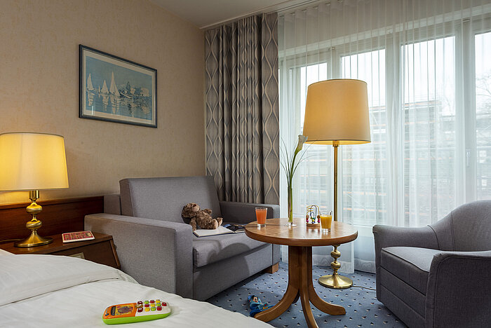 Family room at Maritim Hotel Cologne with toys on the bed and table, cozy seating area, and large windows with curtains.