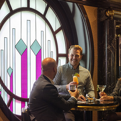 Three guests enjoy cocktails at the piano bar in the Maritim Cologne, sitting in front of a decorative stained glass window.