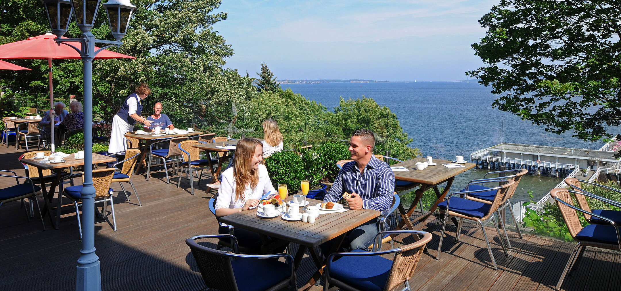 Panorama terrace at the Maritim Hotel Kiel with guests at breakfast, sunny weather and view of the fjord in the background.