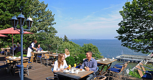 Panorama terrace at the Maritim Hotel Kiel with guests at breakfast, sunny weather and view of the fjord in the background.