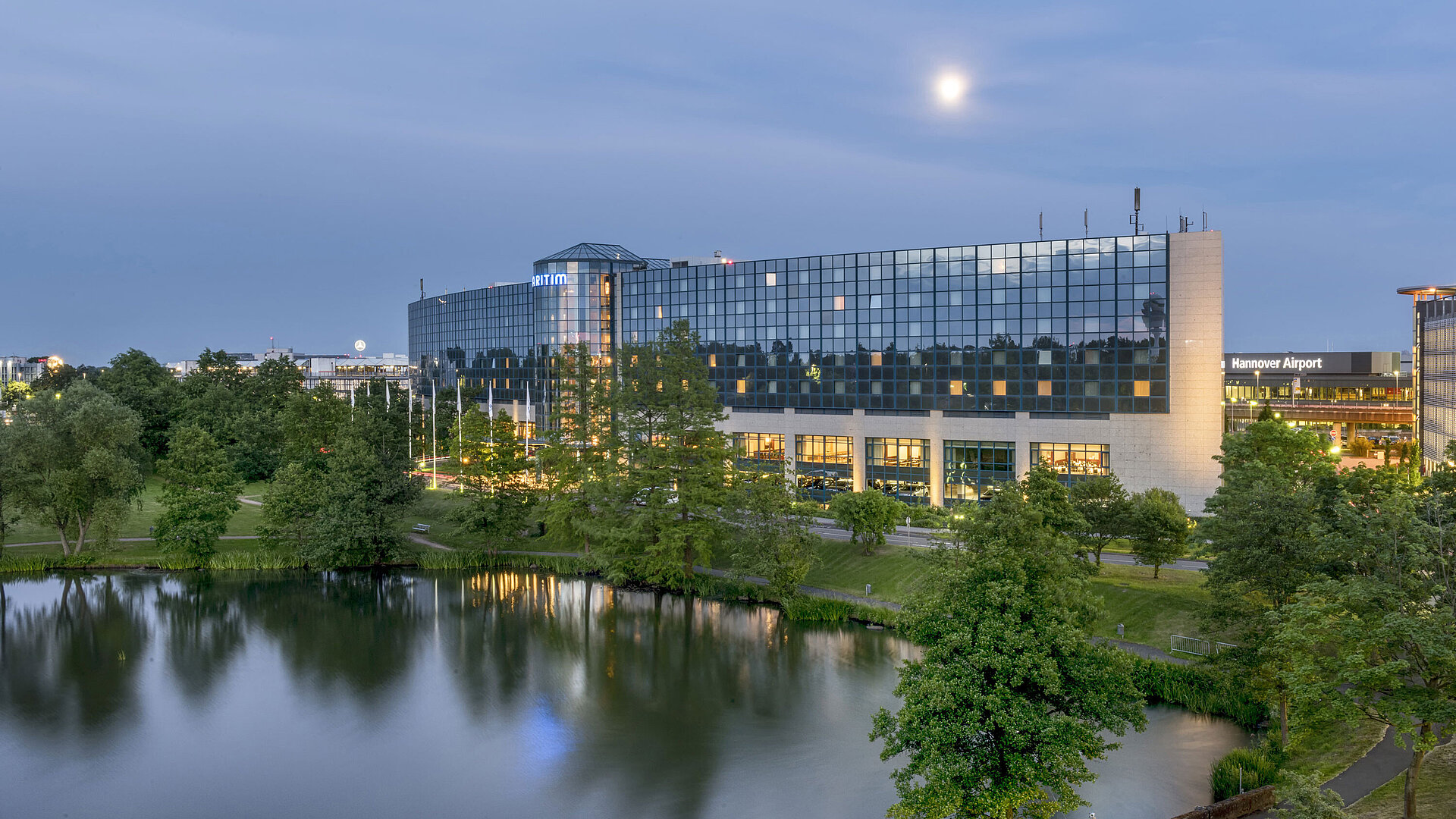 Illuminated Maritim Hotel Hannover with a glass facade and lake reflection at dusk.
