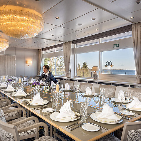 Salon Oslo in the Maritim Hotel Kiel, long festively laid table with sea view, chandelier, staff arranging the room.