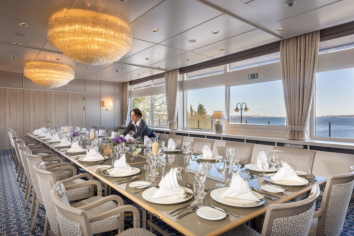 Salon Oslo in the Maritim Hotel Kiel, long festively laid table with sea view, chandelier, staff arranging the room.