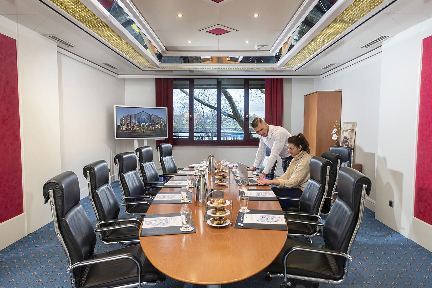 Two people working in the Salon Boardroom at Maritim Cologne with laptops at a conference table with drinks and snacks.