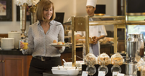 Smiling hotel guest picks fresh food at the breakfast buffet at Maritim Hotel Bad Homburg, chef in the background.